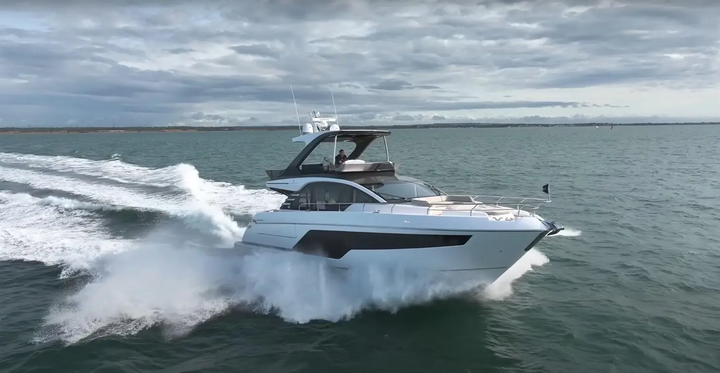 A luxury yacht speeds across blue waters, creating white waves, under a cloudy sky with a person at the helm.